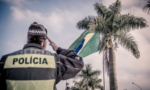 Fotografia de uma pessoa com uma farda da polícia de costas para a câmera batendo continência para a bandeira do Brasil do um coqueiro ao fundo