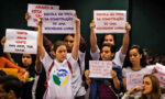 Fotografia mostrando um grupo de jovens, principalmente mulheres, com cartazes, protestando. Em alguns dos cartazes se lê "Escola em prol da construção de uma sociedade livre!"