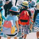 Fotografia de um grupo de crianças com mochilas, chapéus em um dia de sol.