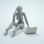 Fotografia de uma boneca cinza sem identidade ao celular, enquanto usa o computador
