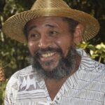 Fotografia do Nego Bispo utilizando um chapéu de palha, em um fundo com plantas
