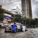 Fotografia de dois homens empurrando um carro preio em meio a uma enchente.