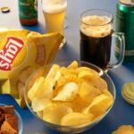 Fotografia de salgadinho de batatas, torradas, azeitonas e cervejas em uma mesa azul.
